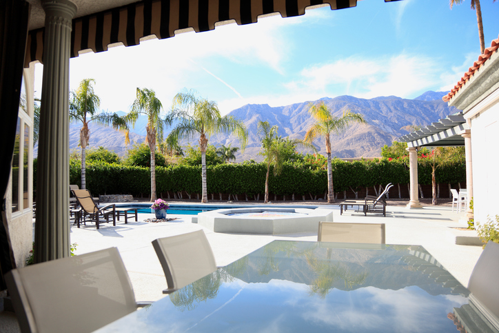 Large pool and outdoor dining area of luxury home. Reflection of mountain range and palm trees in table under cabana. No people. Day time.