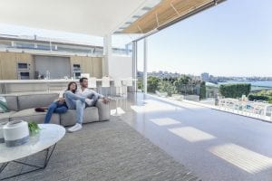 Couple sitting in a modern open plan house. They are sitting on a sofa with a kitchen behind them. They are looking at the beautiful water view from their house. They are attractive, smiling and happy.